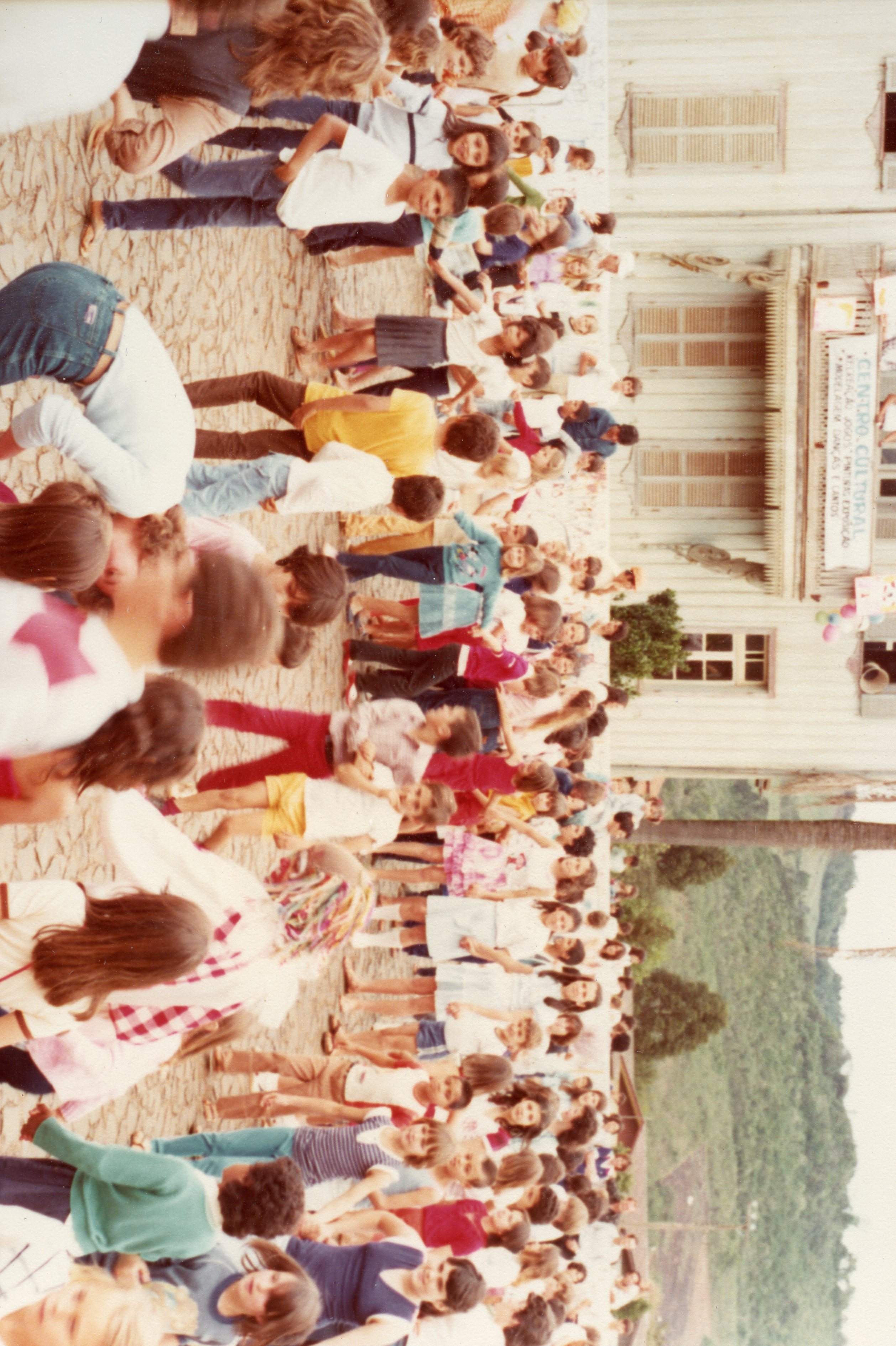 Uma fotografia colorida que captura uma grande aglomeração de pessoas, majoritariamente crianças e jovens, em frente a um edifício de madeira com arquitetura típica de colonização europeia, identificado como 'Centro Cultural'. A cena sugere um evento comunitário ou escolar ao ar livre, com vestimentas que indicam um período entre o final da década de 1970 e início da década de 1980.