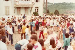 Uma fotografia colorida que captura uma grande aglomeração de pessoas, majoritariamente crianças e jovens, em frente a um edifício de madeira com arquitetura típica de colonização europeia, identificado como 'Centro Cultural'. A cena sugere um evento comunitário ou escolar ao ar livre, com vestimentas que indicam um período entre o final da década de 1970 e início da década de 1980.