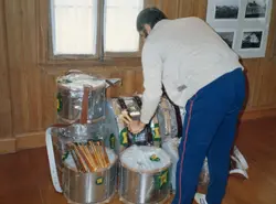 Uma pessoa de costas, vestindo agasalho e calça esportiva, manuseia um conjunto de tambores novos ainda embalados em plástico. O cenário parece ser um ambiente interno com paredes de madeira, possivelmente um museu ou centro cultural, com fotografias antigas expostas ao fundo.
