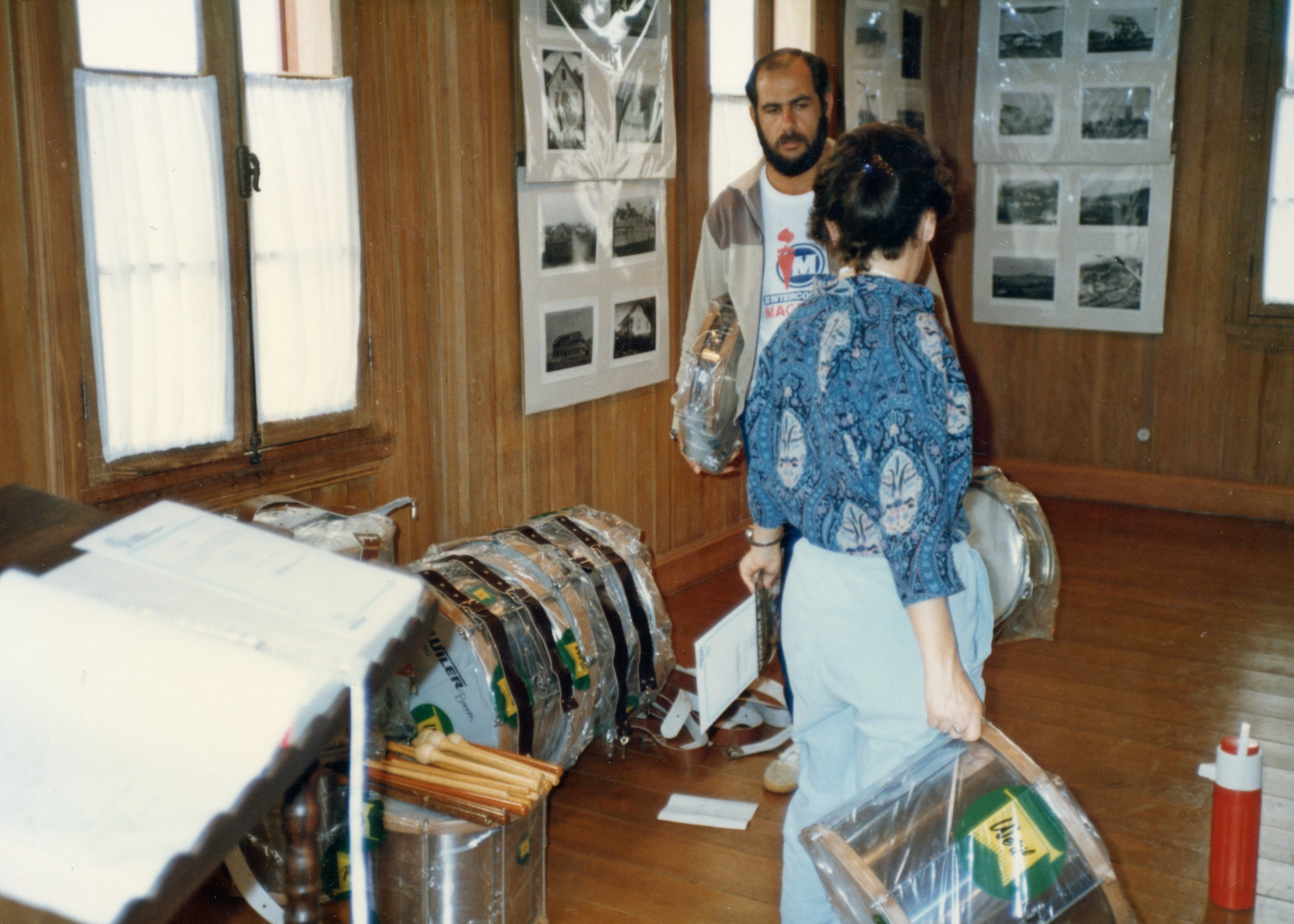 Uma cena interna em um ambiente com paredes de madeira, possivelmente um museu ou centro cultural, onde duas pessoas organizam instrumentos musicais de percussão embalados em plástico. Nas paredes, há exposições de fotografias antigas, sugerindo um contexto de preservação histórica ou organização de um evento cultural.