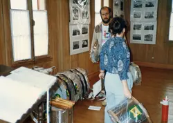 Uma cena interna em um ambiente com paredes de madeira, possivelmente um museu ou centro cultural, onde duas pessoas organizam instrumentos musicais de percussão embalados em plástico. Nas paredes, há exposições de fotografias antigas, sugerindo um contexto de preservação histórica ou organização de um evento cultural.