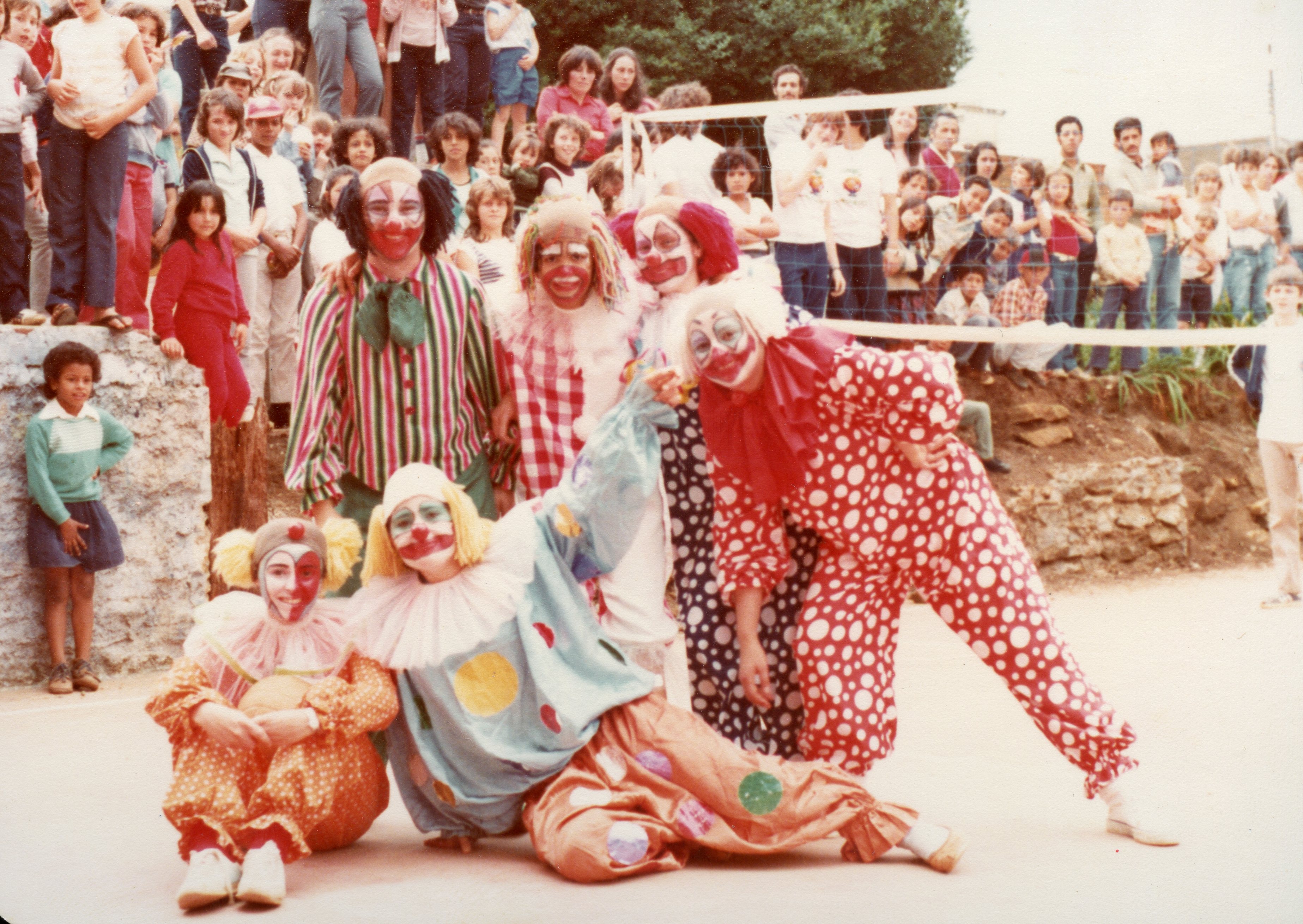Uma fotografia colorida que captura um grupo de pessoas fantasiadas de palhaços posando para uma foto em um ambiente ao ar livre, possivelmente durante um evento comunitário ou escolar. Ao fundo, observa-se uma multidão de espectadores, incluindo muitas crianças, sugerindo uma celebração festiva típica de pequenas cidades brasileiras nas décadas de 1980 ou 1990.