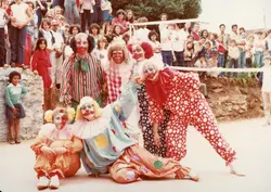 Uma fotografia colorida que captura um grupo de pessoas fantasiadas de palhaços posando para uma foto em um ambiente ao ar livre, possivelmente durante um evento comunitário ou escolar. Ao fundo, observa-se uma multidão de espectadores, incluindo muitas crianças, sugerindo uma celebração festiva típica de pequenas cidades brasileiras nas décadas de 1980 ou 1990.