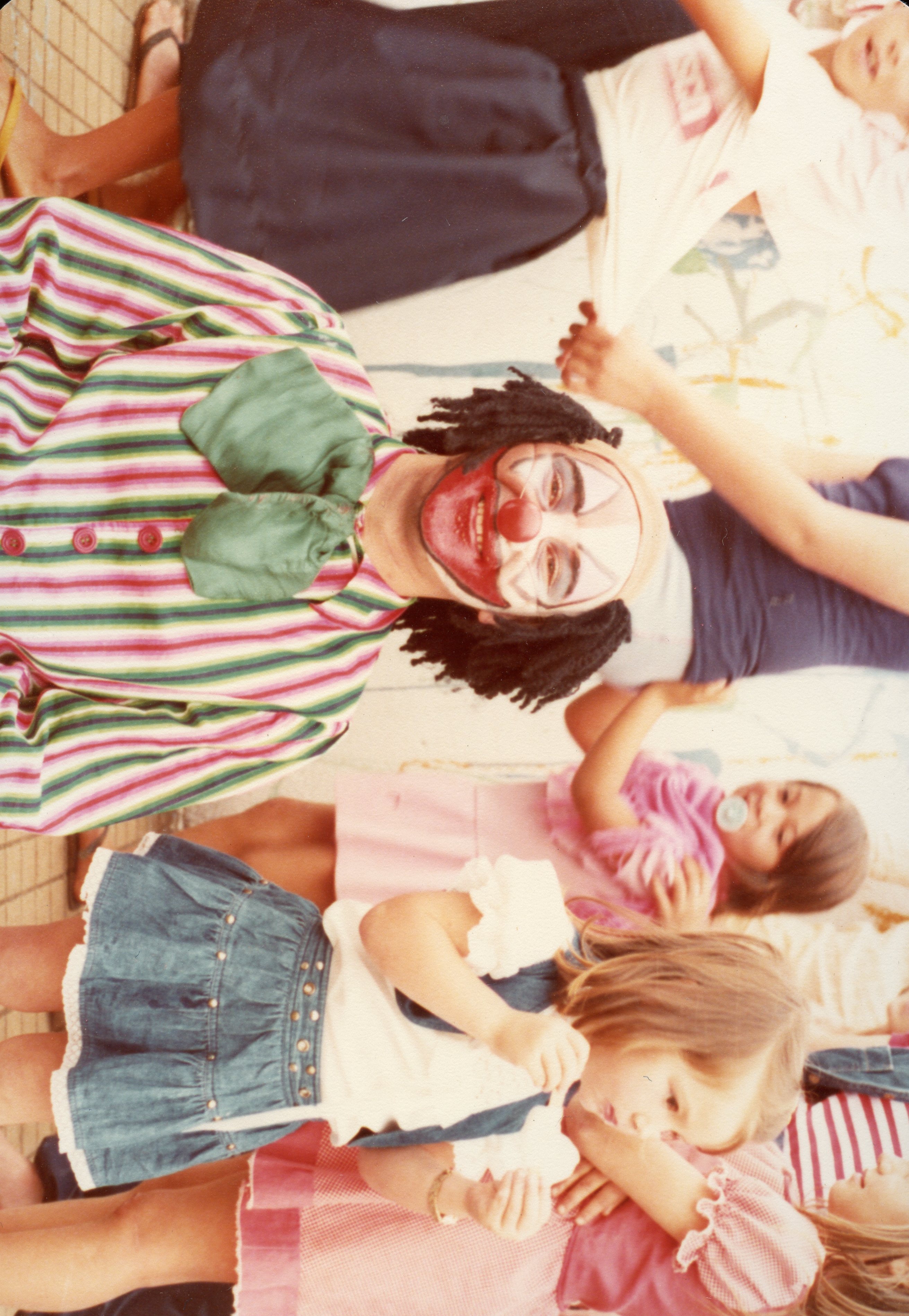 Uma fotografia colorida que captura um momento de lazer com um artista caracterizado como palhaço interagindo com crianças. A imagem apresenta vestimentas típicas das décadas de 1980 ou 1990, sugerindo uma festa infantil ou evento comunitário em um ambiente descontraído.