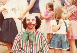 Uma fotografia colorida que captura um momento de lazer com um artista caracterizado como palhaço interagindo com crianças. A imagem apresenta vestimentas típicas das décadas de 1980 ou 1990, sugerindo uma festa infantil ou evento comunitário em um ambiente descontraído.