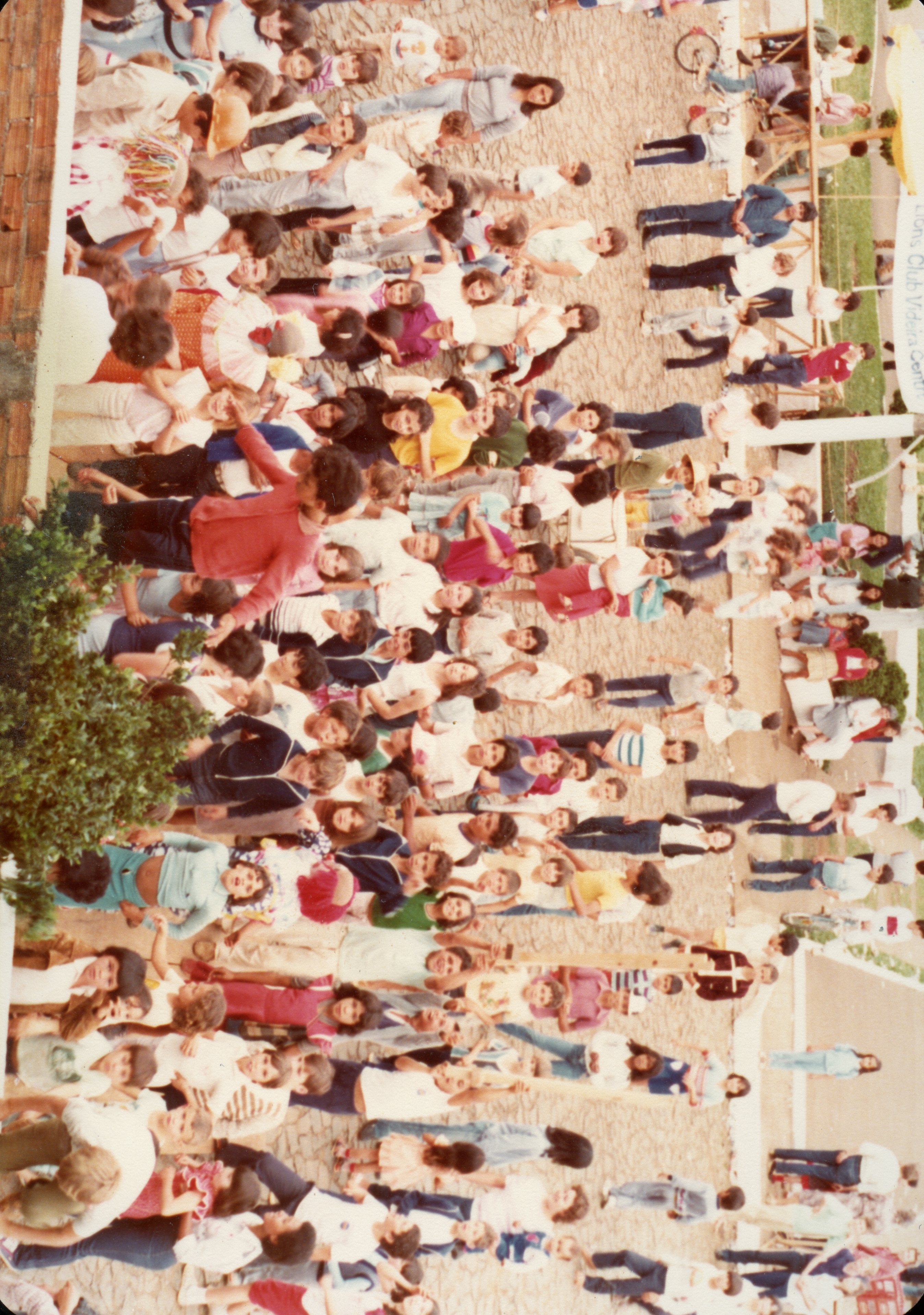 Uma fotografia aérea de uma grande multidão reunida em um espaço aberto, possivelmente em um evento comunitário ou escolar em Videira. As pessoas estão vestidas com roupas casuais típicas das décadas de 1980 ou 1990, e há uma estrutura de madeira visível ao fundo com uma faixa que menciona 'Lions Club'.