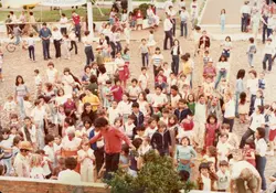 Uma fotografia aérea de uma grande multidão reunida em um espaço aberto, possivelmente em um evento comunitário ou escolar em Videira. As pessoas estão vestidas com roupas casuais típicas das décadas de 1980 ou 1990, e há uma estrutura de madeira visível ao fundo com uma faixa que menciona 'Lions Club'.
