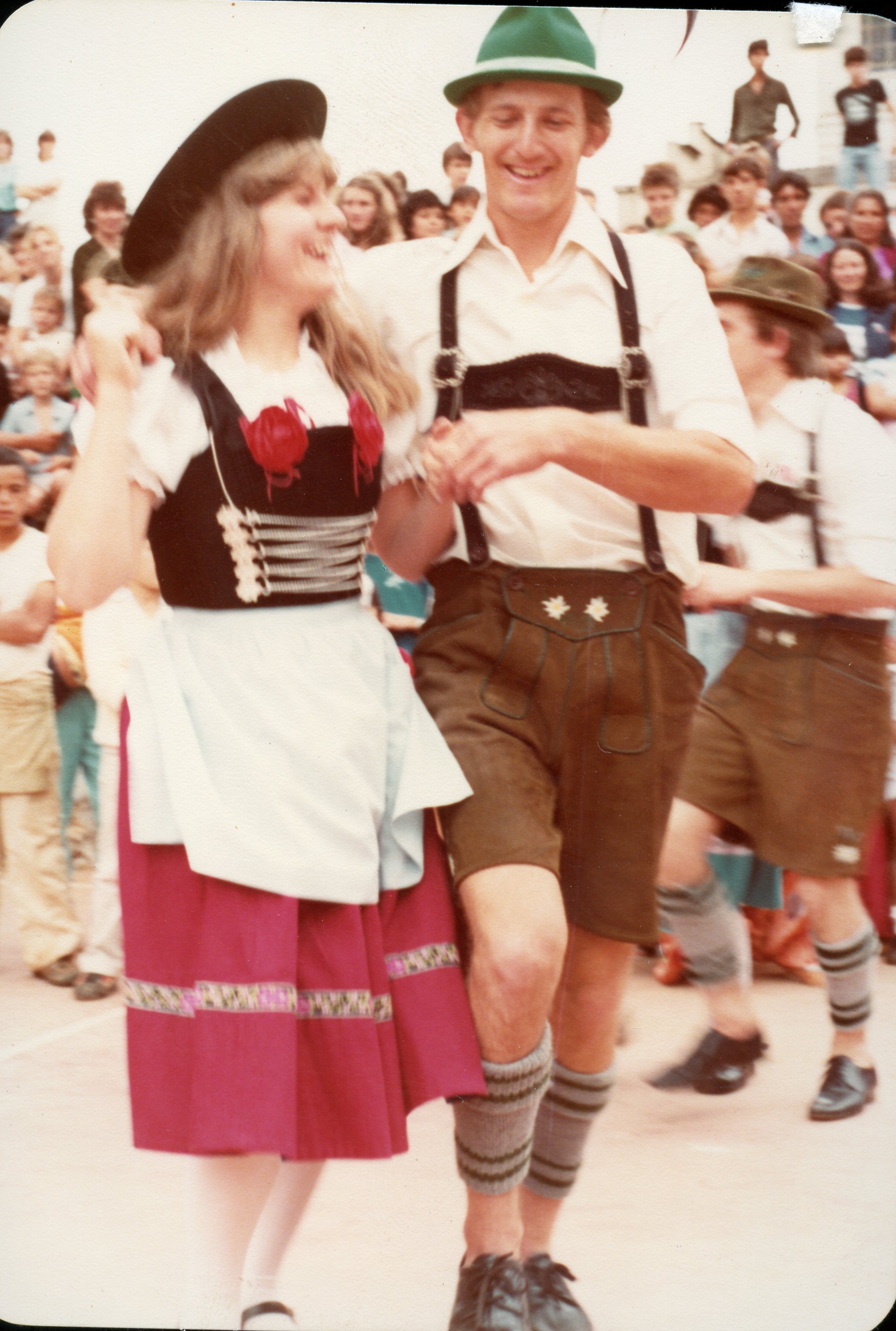 Uma fotografia colorida capturando um casal dançando em um evento cultural, possivelmente uma festa típica alemã em uma comunidade brasileira. Eles vestem trajes tradicionais, como o dirndl e o lederhosen, e estão cercados por uma multidão em um ambiente ao ar livre, sugerindo uma celebração de imigração ou festival folclórico.