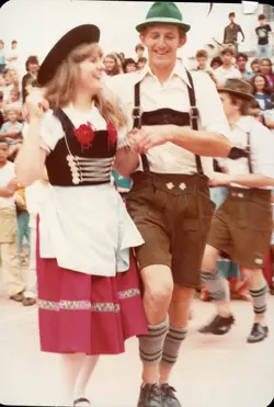 Uma fotografia colorida capturando um casal dançando em um evento cultural, possivelmente uma festa típica alemã em uma comunidade brasileira. Eles vestem trajes tradicionais, como o dirndl e o lederhosen, e estão cercados por uma multidão em um ambiente ao ar livre, sugerindo uma celebração de imigração ou festival folclórico.