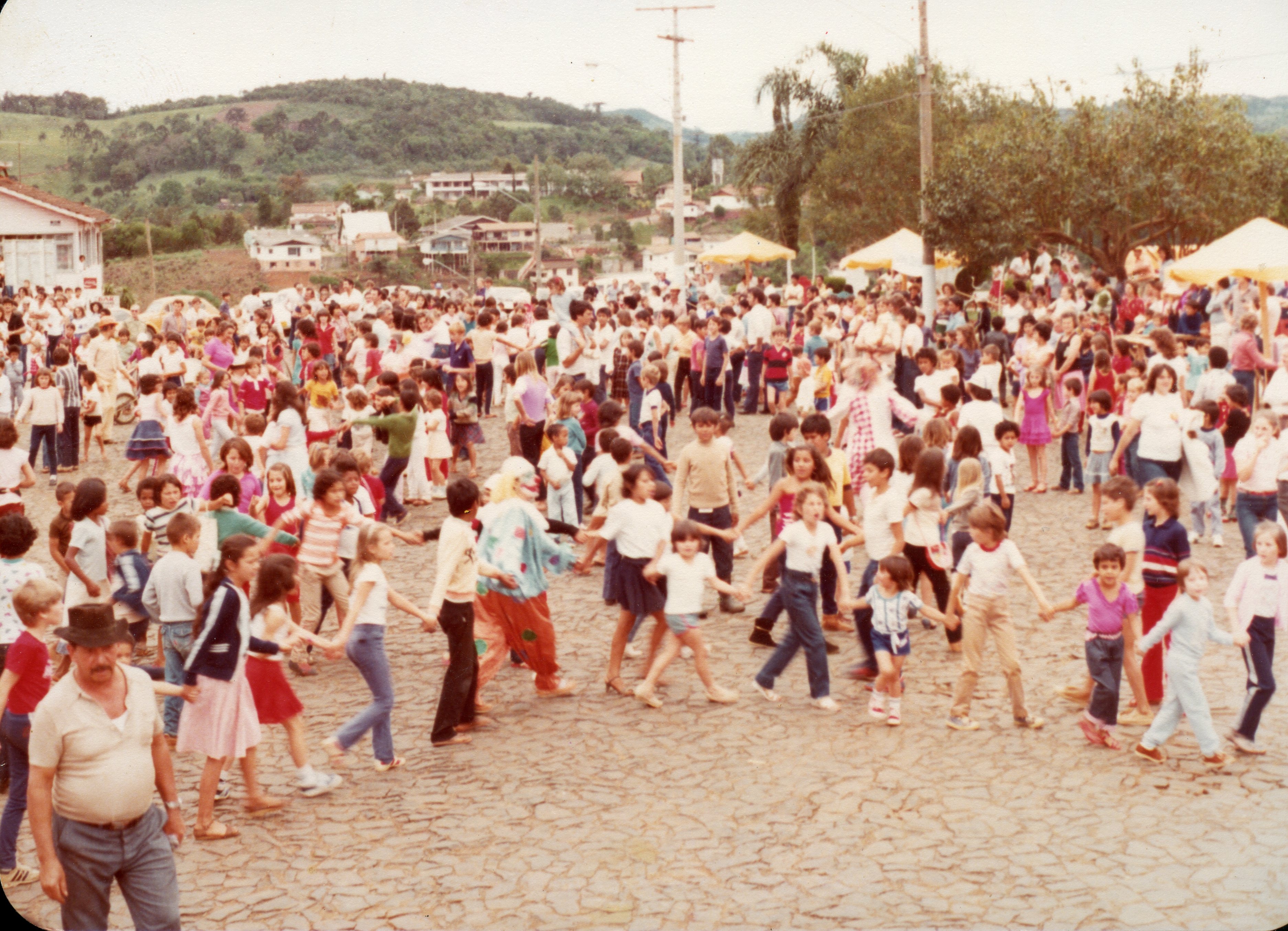 Uma fotografia colorida que retrata uma celebração comunitária ao ar livre, possivelmente uma festa infantil ou evento público em uma pequena cidade brasileira. Crianças e adultos formam uma grande roda em um pátio pavimentado com pedras irregulares, com a presença de um palhaço e barracas ao fundo, sugerindo um contexto festivo típico das décadas de 1970 ou 1980.