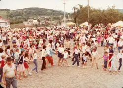 Uma fotografia colorida que retrata uma celebração comunitária ao ar livre, possivelmente uma festa infantil ou evento público em uma pequena cidade brasileira. Crianças e adultos formam uma grande roda em um pátio pavimentado com pedras irregulares, com a presença de um palhaço e barracas ao fundo, sugerindo um contexto festivo típico das décadas de 1970 ou 1980.