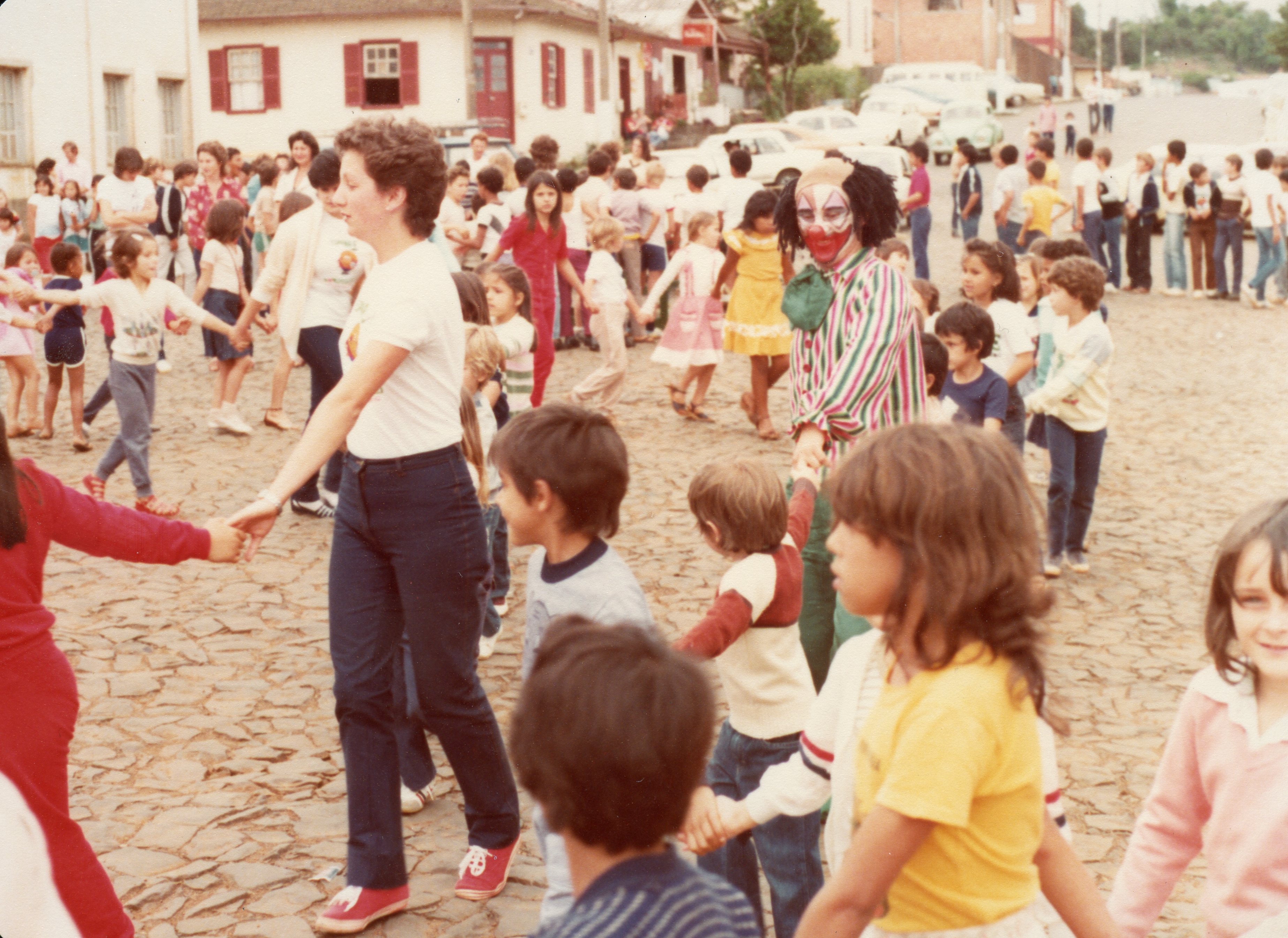 Uma cena festiva ao ar livre em uma rua de paralelepípedos, onde crianças e adultos formam uma grande roda de mãos dadas. Um palhaço caracterizado participa da atividade, sugerindo um evento comunitário ou escolar típico de uma pequena cidade brasileira nas décadas de 1970 ou 1980.
