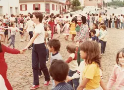 Uma cena festiva ao ar livre em uma rua de paralelepípedos, onde crianças e adultos formam uma grande roda de mãos dadas. Um palhaço caracterizado participa da atividade, sugerindo um evento comunitário ou escolar típico de uma pequena cidade brasileira nas décadas de 1970 ou 1980.