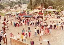Uma cena ao ar livre em uma praça pavimentada com pedras irregulares, mostrando um evento comunitário com muitas crianças e adultos. Ao fundo, vê-se uma paisagem urbana de uma pequena cidade brasileira, possivelmente Videira, com um caminhão de carga e uma faixa indicando 'Centro Cultural'. A vestimenta e a qualidade da fotografia sugerem um evento realizado entre as décadas de 1970 e 1980.