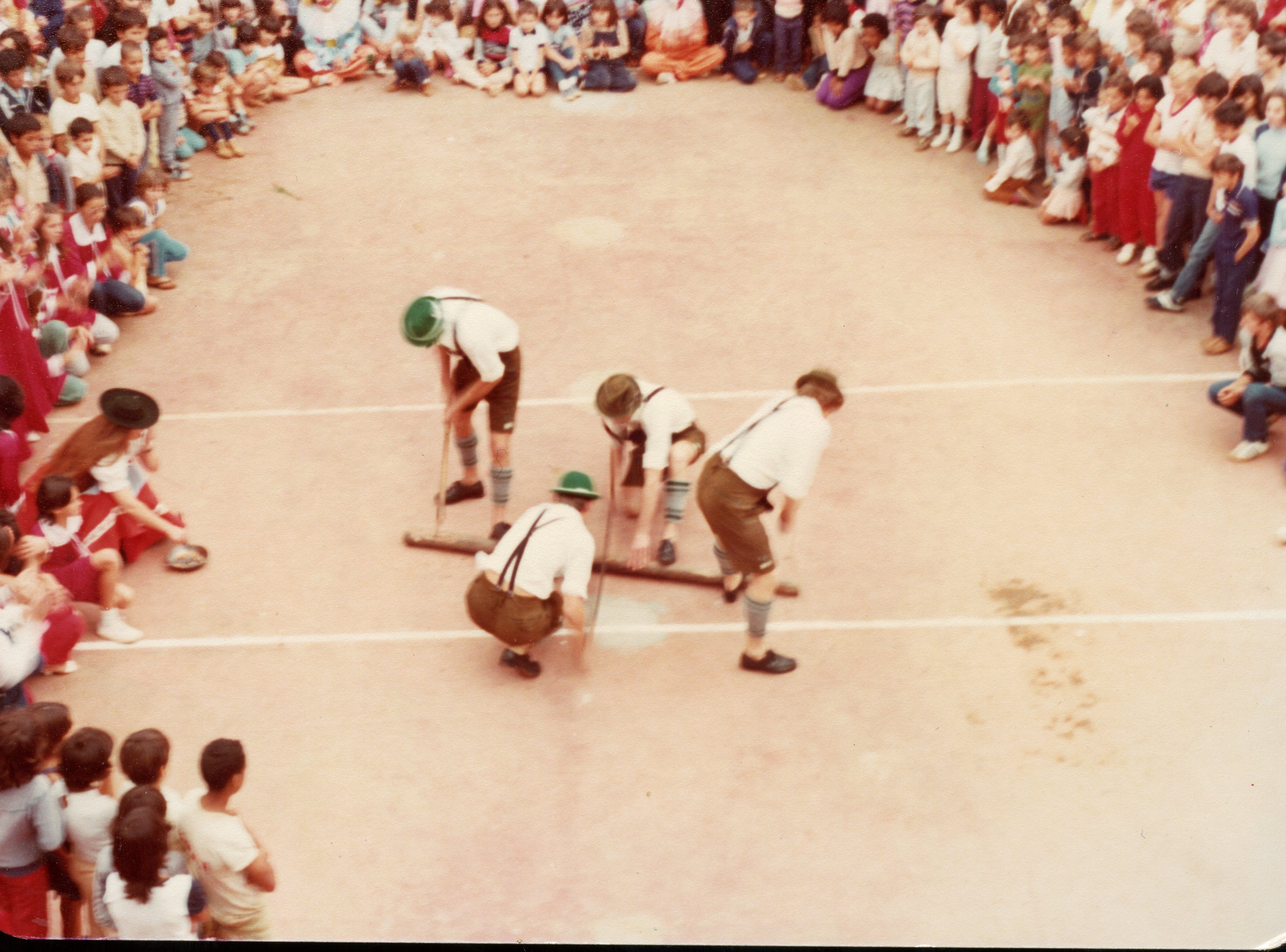 Uma fotografia que captura uma competição ou demonstração cultural, possivelmente uma gincana escolar ou evento comunitário, onde quatro homens vestindo trajes típicos alemães (lederhosen) realizam uma atividade com toras de madeira. O cenário é uma quadra esportiva ao ar livre cercada por uma grande plateia de crianças e jovens, sugerindo um evento escolar ou festividade regional típica do sul do Brasil, provavelmente datada entre as décadas de 1980 e 1990.