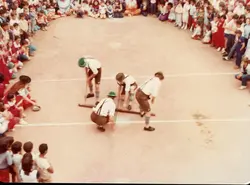 Uma fotografia que captura uma competição ou demonstração cultural, possivelmente uma gincana escolar ou evento comunitário, onde quatro homens vestindo trajes típicos alemães (lederhosen) realizam uma atividade com toras de madeira. O cenário é uma quadra esportiva ao ar livre cercada por uma grande plateia de crianças e jovens, sugerindo um evento escolar ou festividade regional típica do sul do Brasil, provavelmente datada entre as décadas de 1980 e 1990.