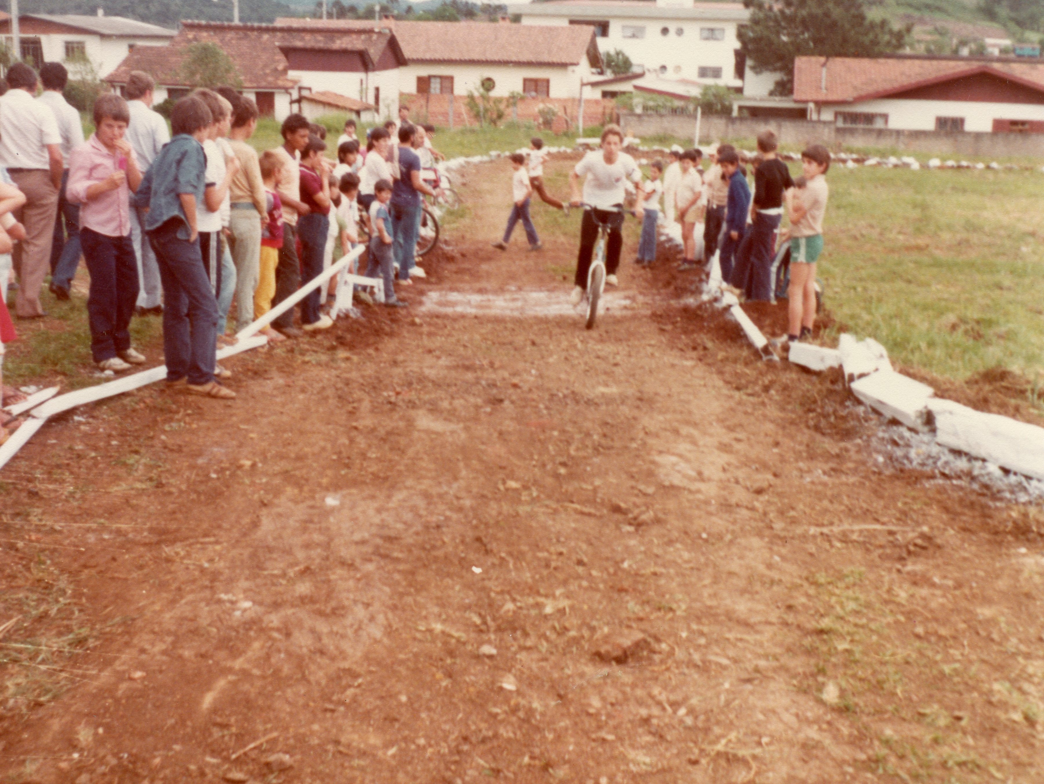Uma fotografia colorida que retrata uma competição de ciclismo amador em uma pista de terra improvisada, provavelmente em uma pequena cidade brasileira. O cenário mostra jovens e adultos observando o evento, com vestimentas típicas da década de 1980, em um ambiente que sugere uma área periférica ou rural em desenvolvimento.