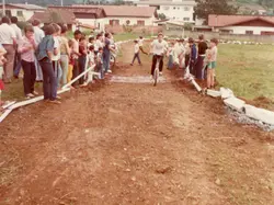 Uma fotografia colorida que retrata uma competição de ciclismo amador em uma pista de terra improvisada, provavelmente em uma pequena cidade brasileira. O cenário mostra jovens e adultos observando o evento, com vestimentas típicas da década de 1980, em um ambiente que sugere uma área periférica ou rural em desenvolvimento.