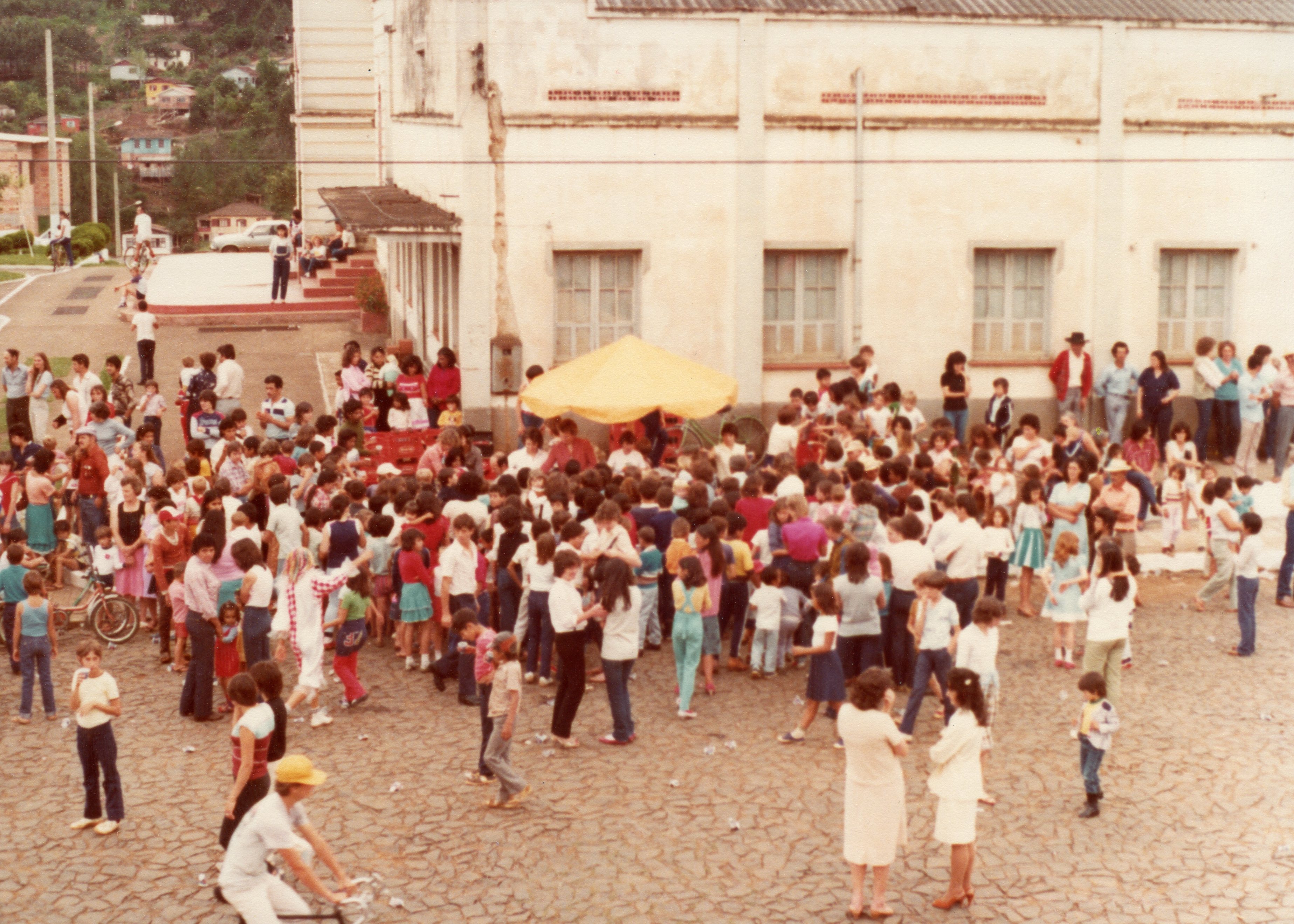 Uma fotografia de época, provavelmente das décadas de 1970 ou 1980, retratando uma grande aglomeração de pessoas em uma praça ou pátio em frente a um edifício institucional. O cenário sugere um evento comunitário ou festividade ao ar livre, com a presença de um guarda-sol amarelo, vestimentas casuais da época e um calçamento de pedras irregulares.