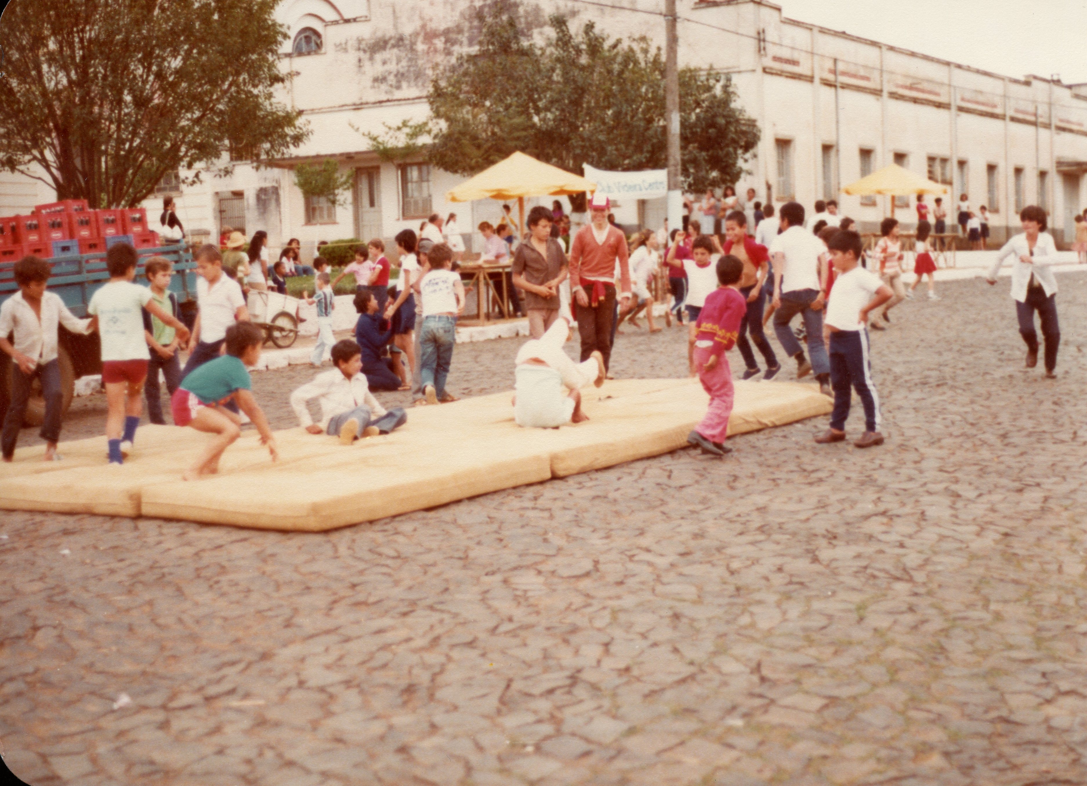 Uma cena ao ar livre em uma praça pavimentada com paralelepípedos, onde crianças e jovens se reúnem para atividades recreativas sobre colchões dispostos no chão. O ambiente sugere um evento comunitário ou escolar em uma pequena cidade brasileira, com arquitetura de época ao fundo e vestimentas típicas das décadas de 1980 ou 1990.