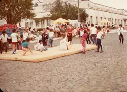 Uma cena ao ar livre em uma praça pavimentada com paralelepípedos, onde crianças e jovens se reúnem para atividades recreativas sobre colchões dispostos no chão. O ambiente sugere um evento comunitário ou escolar em uma pequena cidade brasileira, com arquitetura de época ao fundo e vestimentas típicas das décadas de 1980 ou 1990.