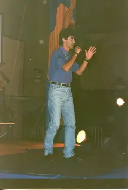 Uma fotografia de um homem em um palco, segurando um microfone e gesticulando, possivelmente durante uma apresentação ou palestra. O estilo das roupas, como a camisa polo e o jeans de corte clássico, sugere um contexto brasileiro das décadas de 1980 ou 1990.