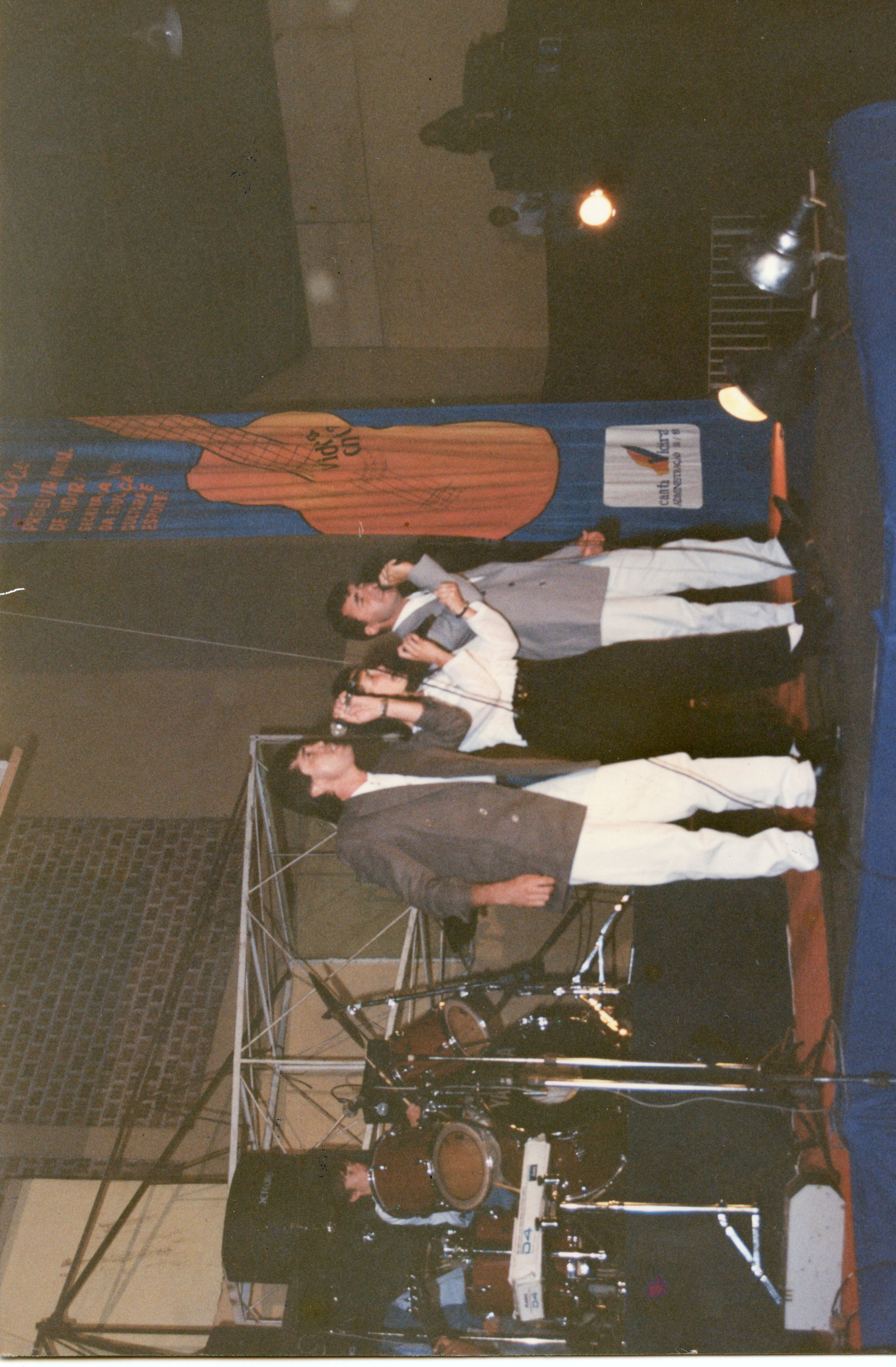 Uma fotografia colorida de um evento musical em um ambiente interno, possivelmente um ginásio ou centro cultural em Videira. Três músicos aparecem no palco cantando com microfones, acompanhados por uma bateria, com um banner promocional ao fundo que sugere um festival ou evento de cultura e esporte.