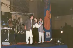 Uma fotografia colorida de um evento musical em um ambiente interno, possivelmente um ginásio ou centro cultural em Videira. Três músicos aparecem no palco cantando com microfones, acompanhados por uma bateria, com um banner promocional ao fundo que sugere um festival ou evento de cultura e esporte.