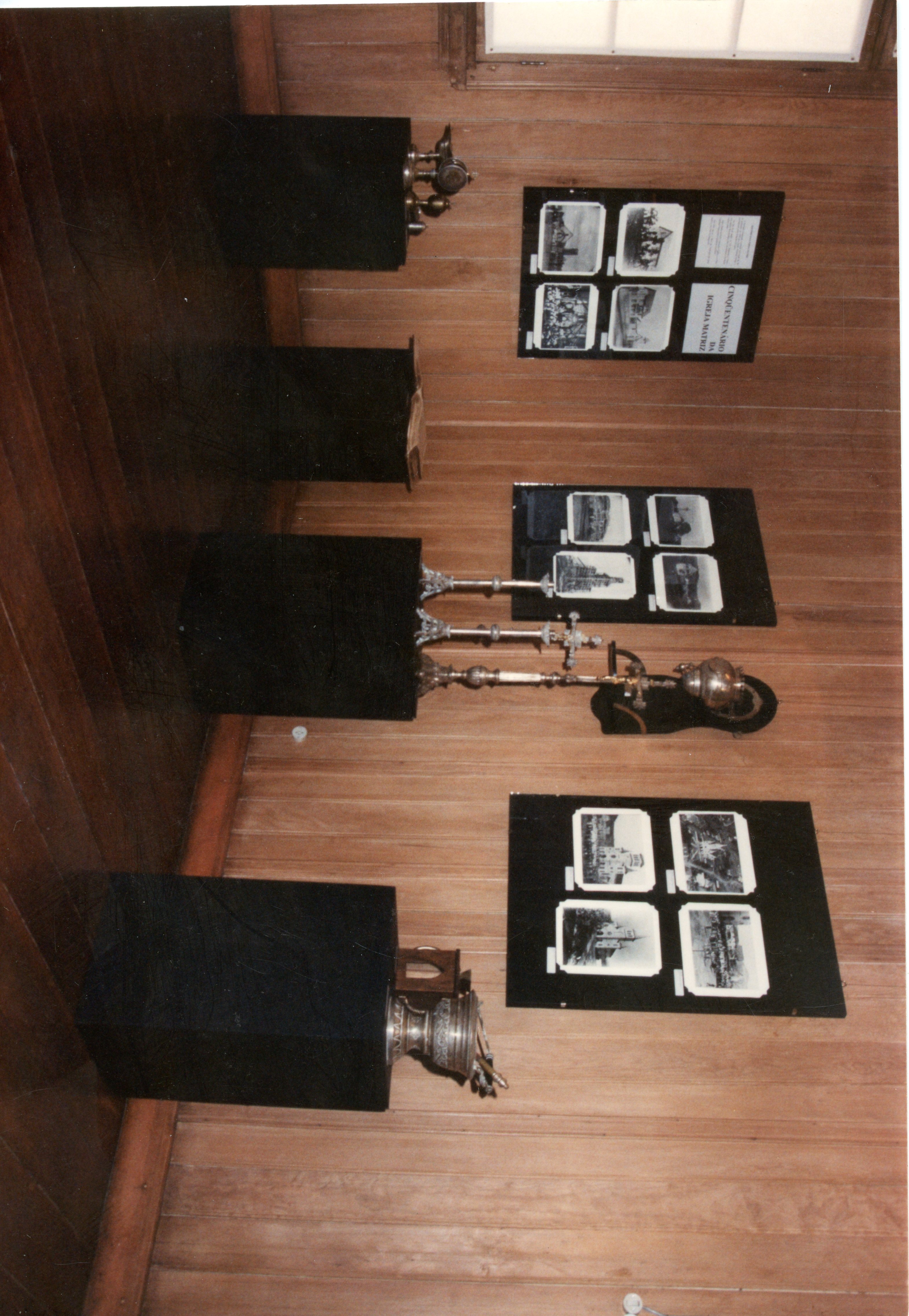 A imagem mostra uma exposição museológica em um ambiente com paredes de madeira, contendo painéis informativos com fotografias históricas e objetos litúrgicos sobre pedestais pretos. O contexto sugere uma mostra comemorativa, possivelmente relacionada ao cinquentenário de uma igreja matriz, comum em registros de memória local em cidades brasileiras.