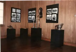 A imagem mostra uma exposição museológica em um ambiente com paredes de madeira, contendo painéis informativos com fotografias históricas e objetos litúrgicos sobre pedestais pretos. O contexto sugere uma mostra comemorativa, possivelmente relacionada ao cinquentenário de uma igreja matriz, comum em registros de memória local em cidades brasileiras.