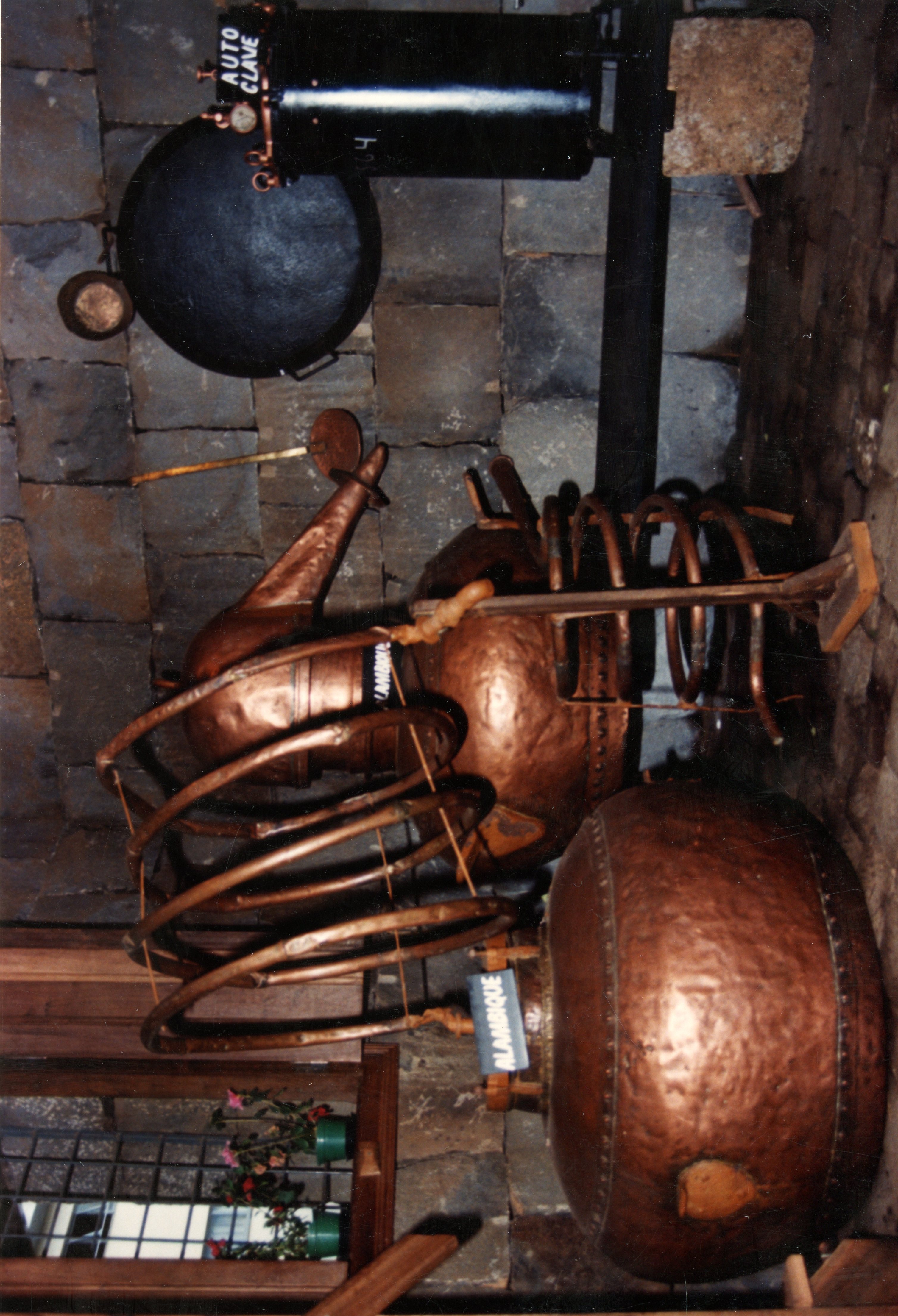 A imagem exibe um alambique de cobre tradicional utilizado para a destilação de bebidas, possivelmente cachaça, em um ambiente rústico com paredes de pedra. O equipamento apresenta serpentinas de cobre e uma estrutura robusta, indicando uma prática artesanal comum na colonização rural brasileira.