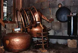 A imagem exibe um alambique de cobre tradicional utilizado para a destilação de bebidas, possivelmente cachaça, em um ambiente rústico com paredes de pedra. O equipamento apresenta serpentinas de cobre e uma estrutura robusta, indicando uma prática artesanal comum na colonização rural brasileira.