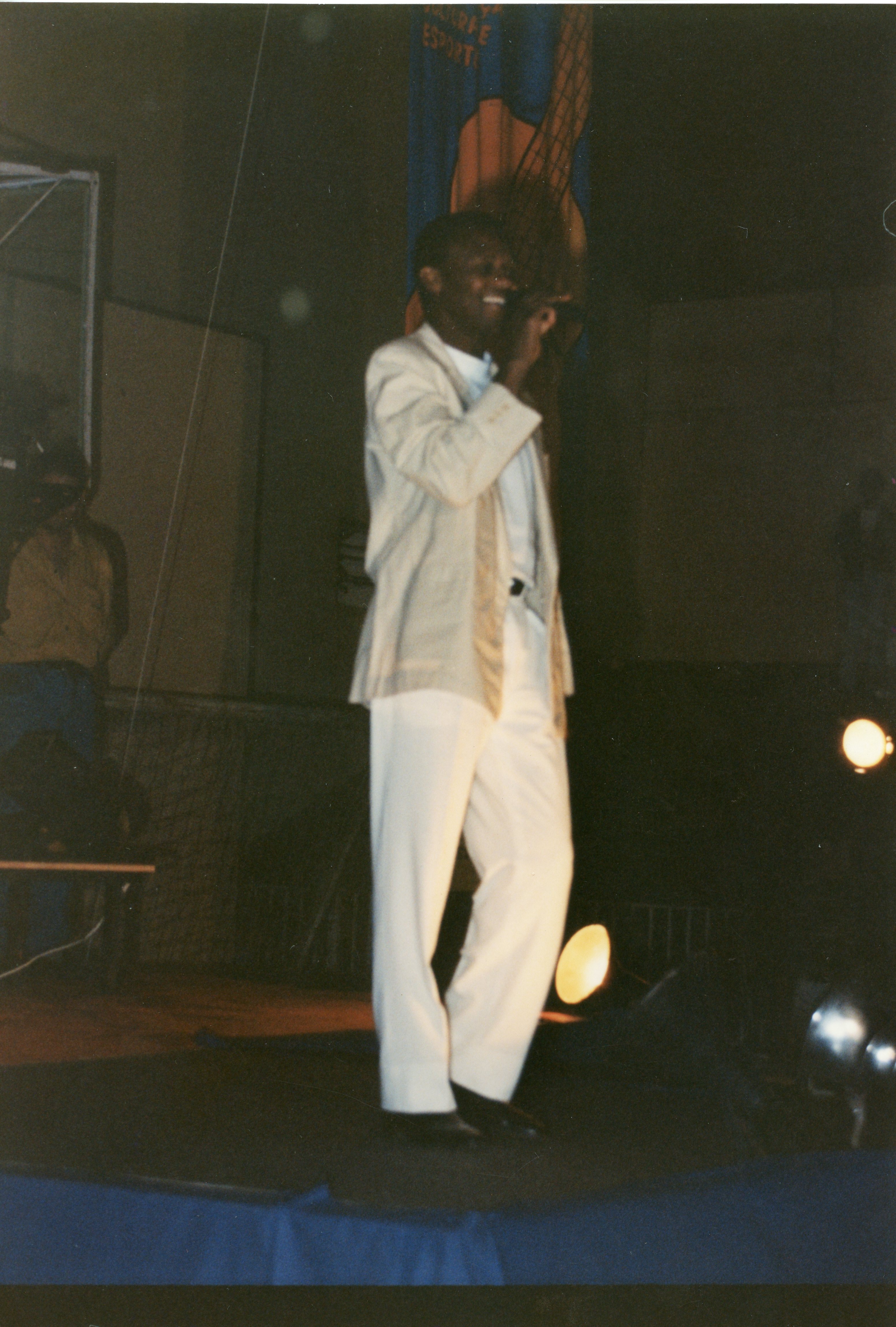 Uma fotografia colorida de um homem negro cantando em um palco, vestindo um terno claro e calças brancas. O ambiente sugere um evento público ou apresentação cultural, possivelmente em um ginásio ou auditório, com iluminação cênica focada no artista.