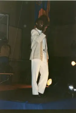 Uma fotografia colorida de um homem negro cantando em um palco, vestindo um terno claro e calças brancas. O ambiente sugere um evento público ou apresentação cultural, possivelmente em um ginásio ou auditório, com iluminação cênica focada no artista.