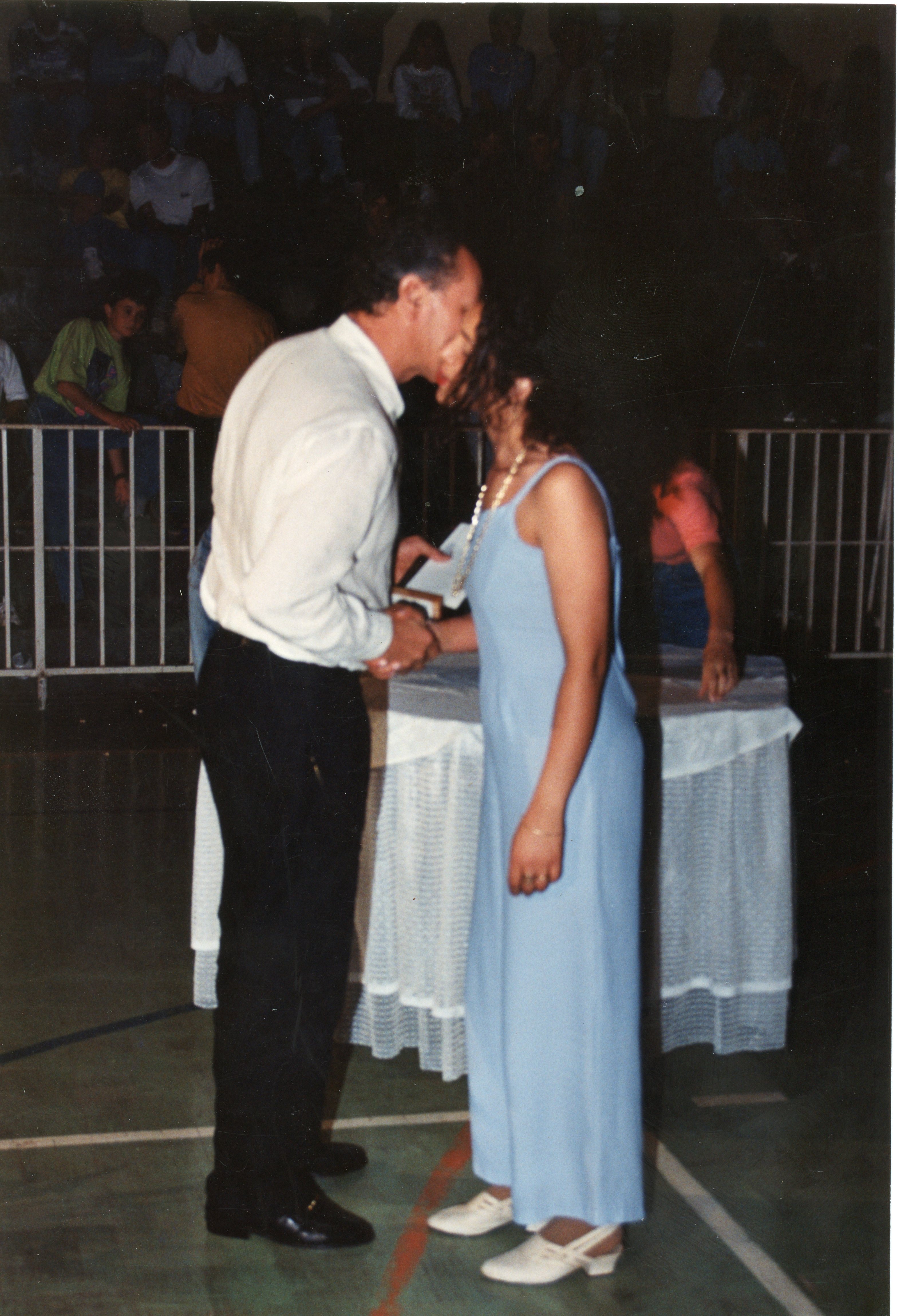 Uma fotografia que registra um momento de entrega de diploma ou premiação em um ambiente interno, possivelmente um ginásio escolar. Um homem de camisa clara e calça escura cumprimenta uma mulher vestindo um longo vestido azul claro, com uma plateia ao fundo, sugerindo um evento formal de formatura ou celebração comunitária típico das décadas de 1980 ou 1990.