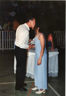 Uma fotografia que registra um momento de entrega de diploma ou premiação em um ambiente interno, possivelmente um ginásio escolar. Um homem de camisa clara e calça escura cumprimenta uma mulher vestindo um longo vestido azul claro, com uma plateia ao fundo, sugerindo um evento formal de formatura ou celebração comunitária típico das décadas de 1980 ou 1990.