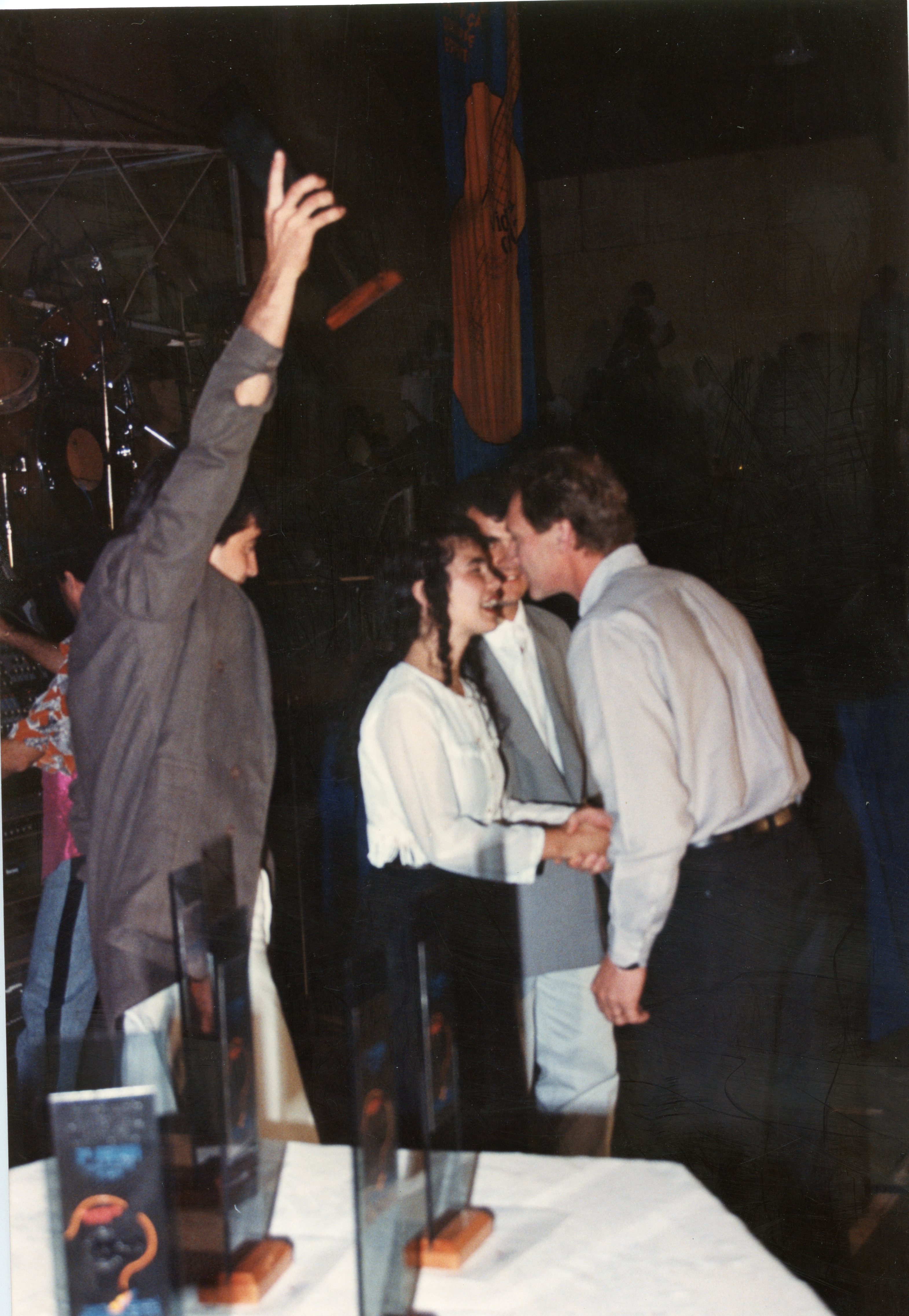 Uma fotografia que captura um momento de celebração ou premiação em um ambiente interno, possivelmente um evento social ou corporativo. Pessoas vestidas com trajes formais da década de 1990 interagem enquanto uma pessoa ao fundo levanta a mão, sugerindo um clima de entusiasmo ou reconhecimento.