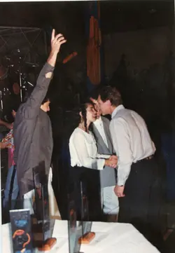 Uma fotografia que captura um momento de celebração ou premiação em um ambiente interno, possivelmente um evento social ou corporativo. Pessoas vestidas com trajes formais da década de 1990 interagem enquanto uma pessoa ao fundo levanta a mão, sugerindo um clima de entusiasmo ou reconhecimento.
