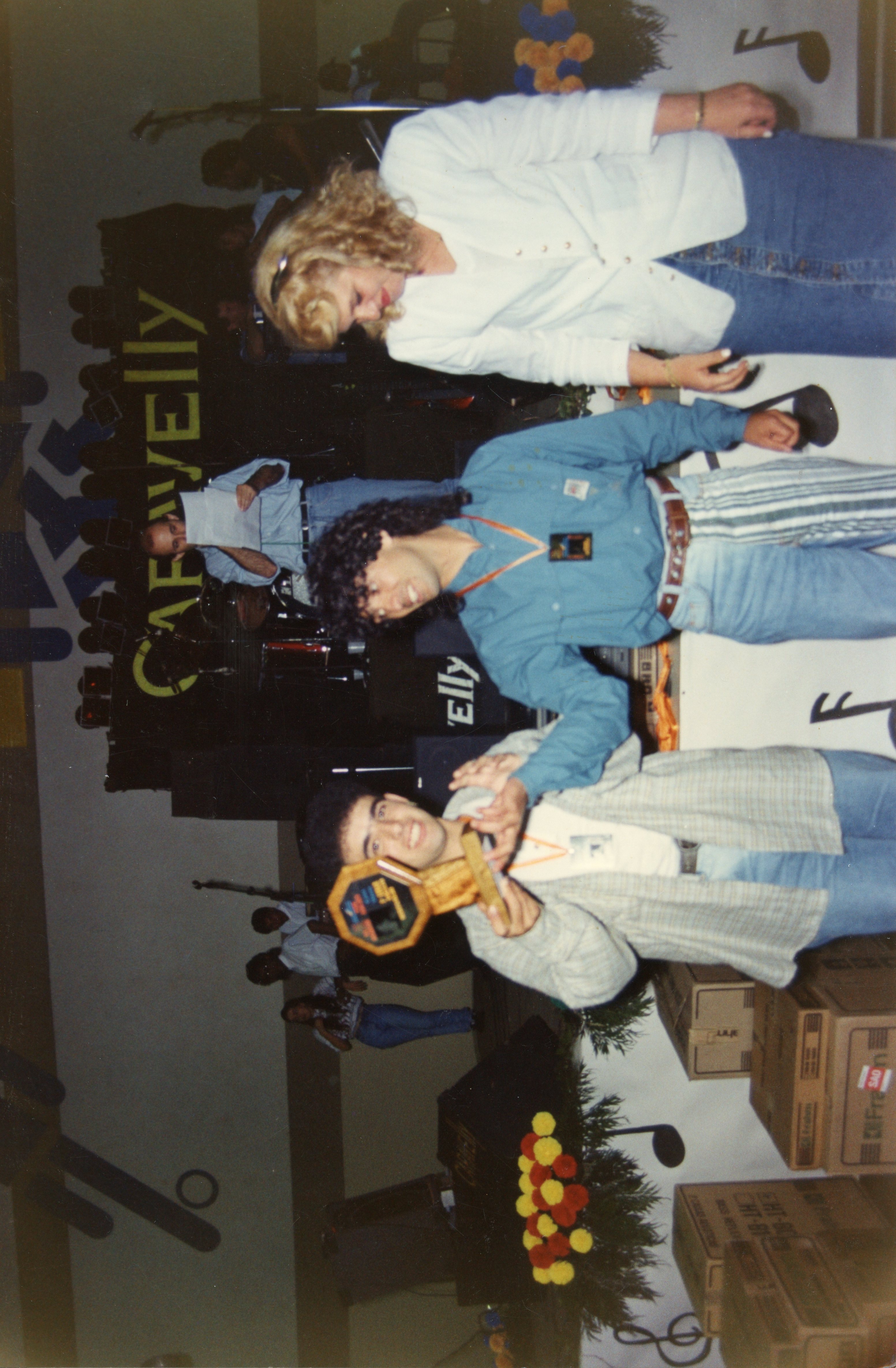 Uma fotografia de um evento social ou premiação em ambiente interno, possivelmente na década de 1990, com pessoas segurando um troféu. O cenário apresenta decoração com notas musicais e o nome 'Carvelly', sugerindo um contexto de festival ou celebração musical.