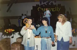 Uma fotografia de um evento social ou premiação em ambiente interno, possivelmente na década de 1990, com pessoas segurando um troféu. O cenário apresenta decoração com notas musicais e o nome 'Carvelly', sugerindo um contexto de festival ou celebração musical.