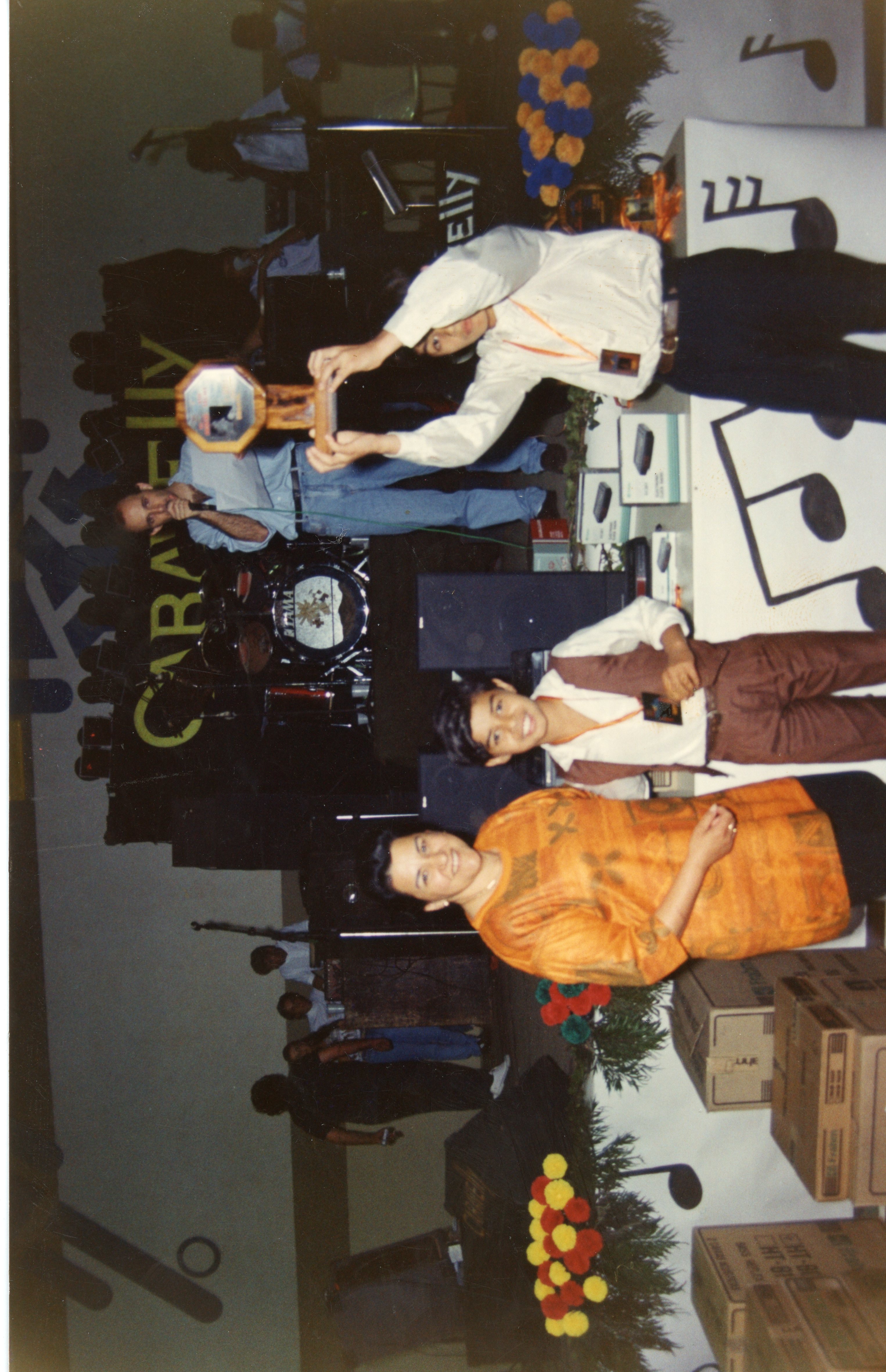 Uma fotografia de um evento interno, possivelmente uma premiação ou celebração escolar, com pessoas posando e segurando um troféu. O ambiente apresenta decoração temática musical, equipamentos de som e caixas de papelão, sugerindo um contexto de organização de eventos ou feira em uma cidade brasileira, provavelmente na década de 1990.