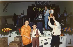 Uma fotografia de um evento interno, possivelmente uma premiação ou celebração escolar, com pessoas posando e segurando um troféu. O ambiente apresenta decoração temática musical, equipamentos de som e caixas de papelão, sugerindo um contexto de organização de eventos ou feira em uma cidade brasileira, provavelmente na década de 1990.