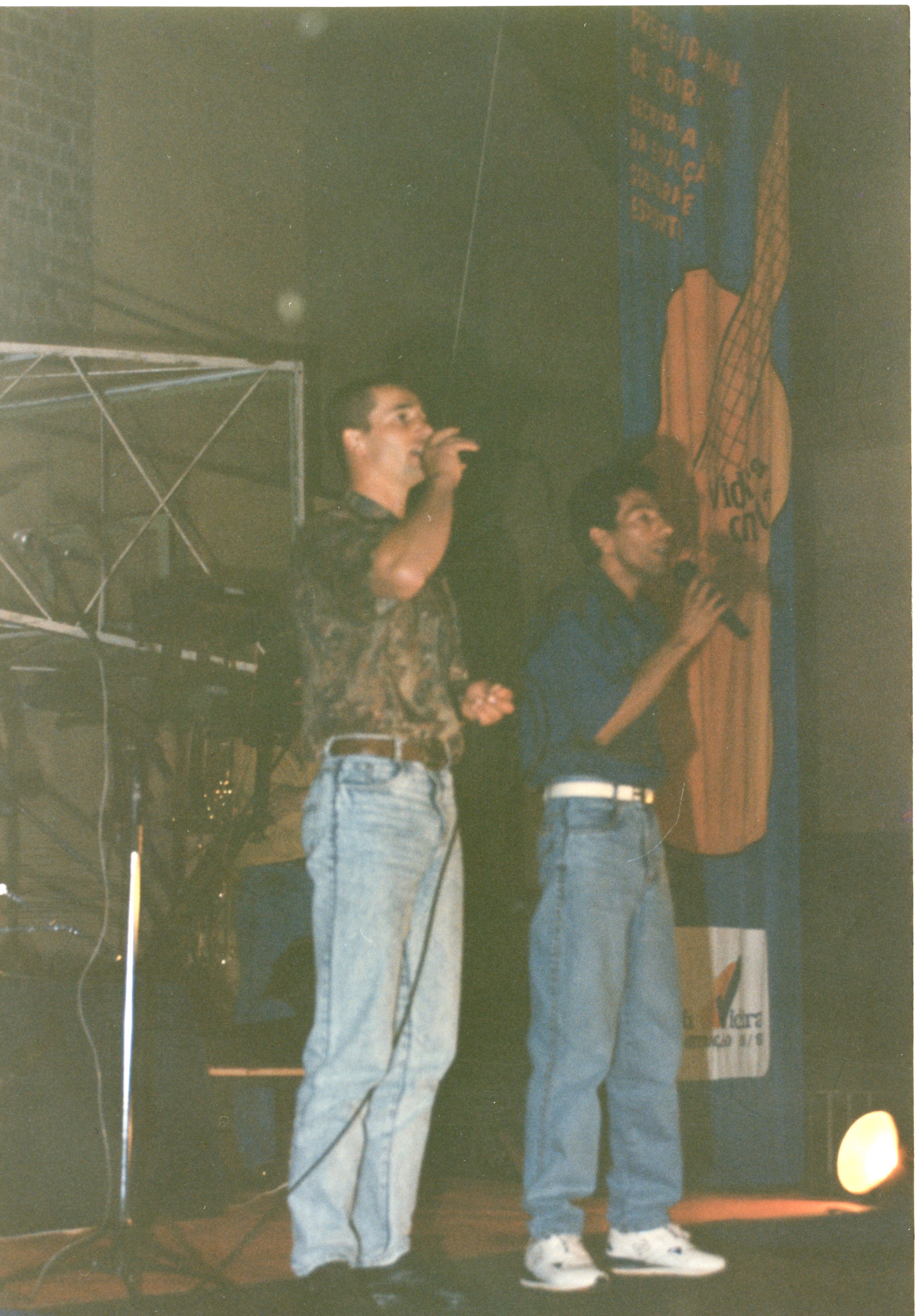 Dois homens cantam em um palco, segurando microfones, em um evento que parece ser uma apresentação musical ou cultural. O cenário inclui um banner vertical com ilustrações e textos, sugerindo um contexto de celebração ou evento público, possivelmente em uma cidade como Videira, com vestimentas típicas da década de 1990.