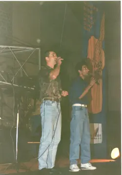 Dois homens cantam em um palco, segurando microfones, em um evento que parece ser uma apresentação musical ou cultural. O cenário inclui um banner vertical com ilustrações e textos, sugerindo um contexto de celebração ou evento público, possivelmente em uma cidade como Videira, com vestimentas típicas da década de 1990.