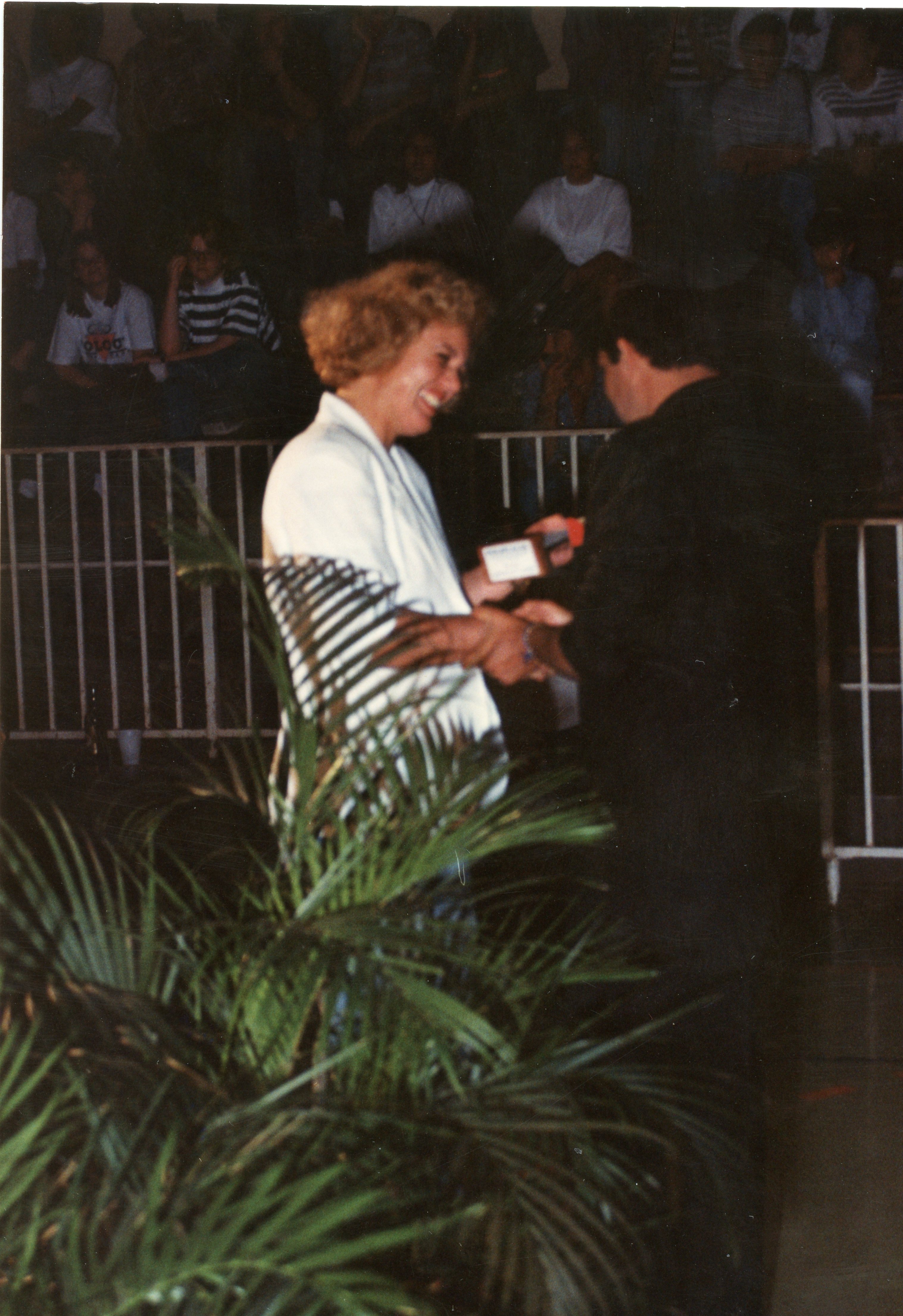 Uma fotografia que captura um momento de entrega de premiação ou homenagem em um ambiente interno, possivelmente um ginásio ou auditório. Uma mulher de cabelos claros e blazer branco sorri ao receber um objeto de um homem, com uma plateia desfocada ao fundo e uma planta em primeiro plano, sugerindo um evento social ou comunitário típico das décadas de 1980 ou 1990.