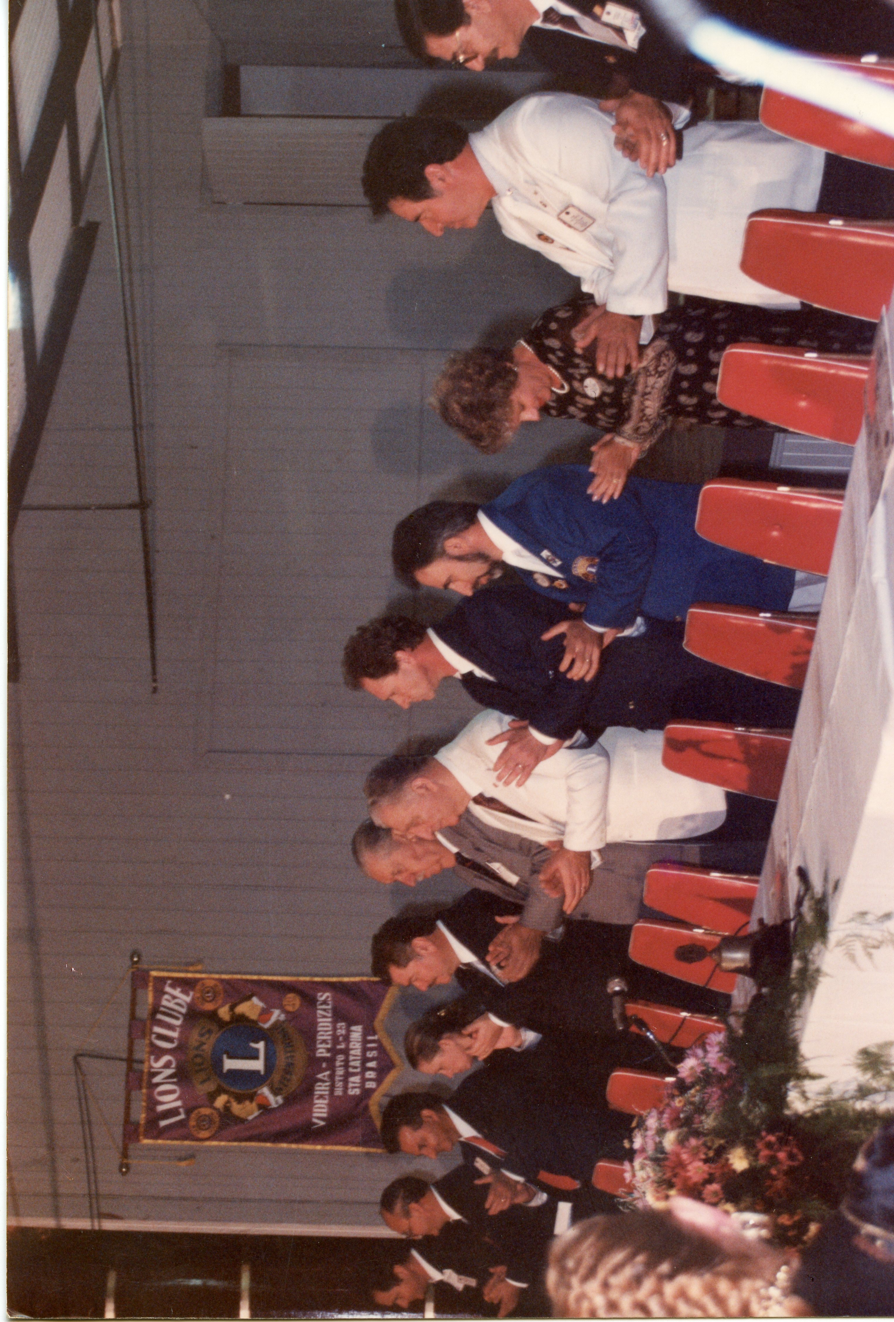 Uma fotografia de um evento formal do Lions Clube em Videira, Santa Catarina, mostrando um grupo de pessoas em fila, possivelmente durante uma cerimônia de posse ou homenagem. Os participantes vestem trajes sociais e formais, típicos de eventos associativos das décadas de 1980 ou 1990, com um estandarte do clube visível ao fundo.