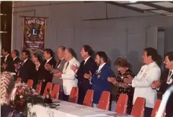 Uma fotografia de um evento formal do Lions Clube em Videira, Santa Catarina, mostrando um grupo de pessoas em fila, possivelmente durante uma cerimônia de posse ou homenagem. Os participantes vestem trajes sociais e formais, típicos de eventos associativos das décadas de 1980 ou 1990, com um estandarte do clube visível ao fundo.
