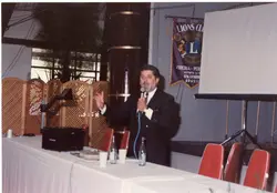 Uma fotografia de um homem de terno discursando em um evento do Lions Clube em Videira, Santa Catarina. O ambiente parece ser um salão de eventos com uma bandeira do clube visível e equipamentos de projeção, sugerindo uma reunião formal ou cerimônia nas décadas de 1980 ou 1990.