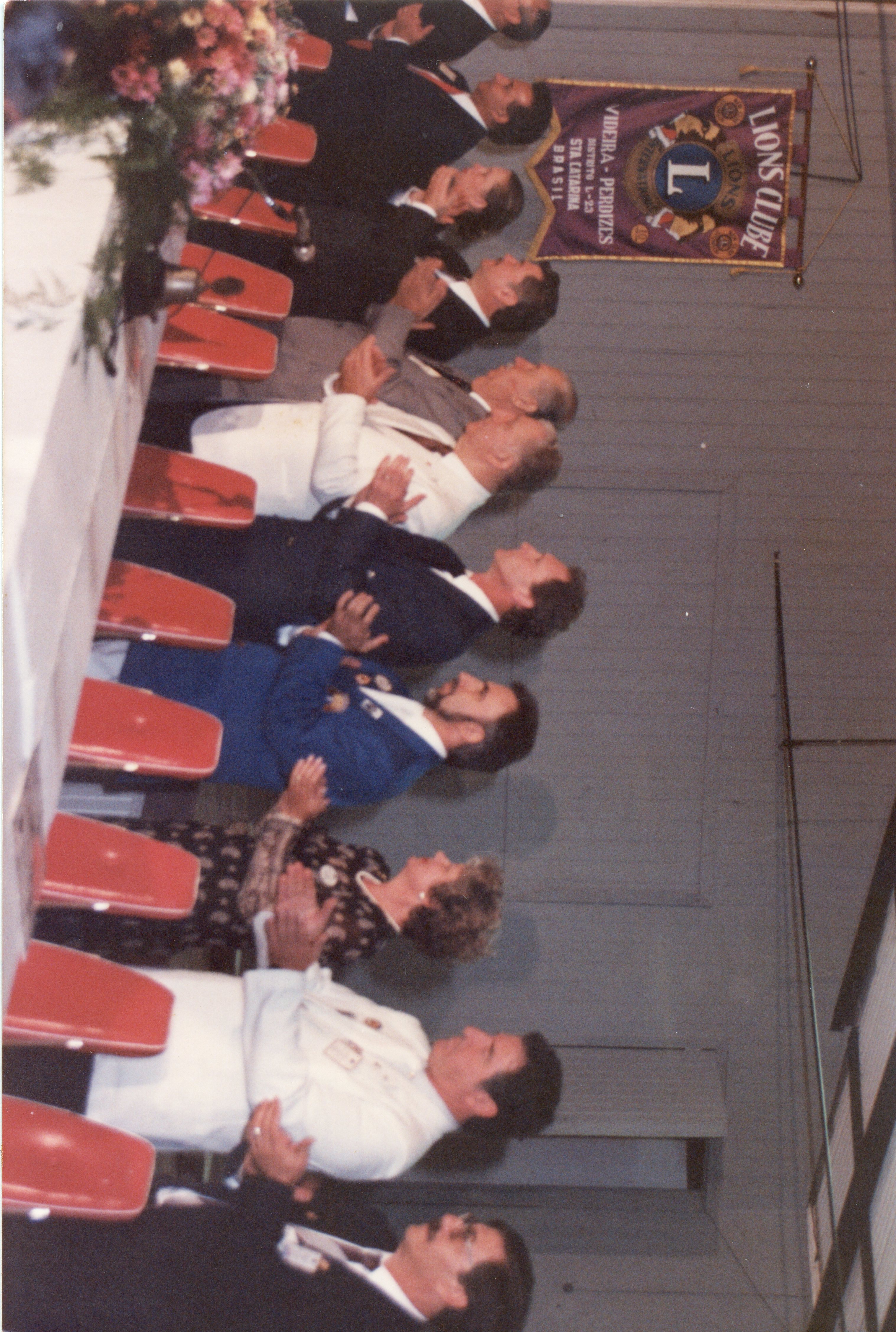 Uma fotografia de um evento formal do Lions Clube em Videira, Santa Catarina, mostrando um grupo de pessoas sentadas à mesa aplaudindo. O ambiente é um salão interno com decoração institucional, incluindo um estandarte do Lions Clube, sugerindo uma reunião de associados ou cerimônia oficial nas décadas de 1980 ou 1990.