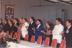 Uma fotografia de um evento formal do Lions Clube em Videira, Santa Catarina, mostrando um grupo de pessoas sentadas à mesa aplaudindo. O ambiente é um salão interno com decoração institucional, incluindo um estandarte do Lions Clube, sugerindo uma reunião de associados ou cerimônia oficial nas décadas de 1980 ou 1990.